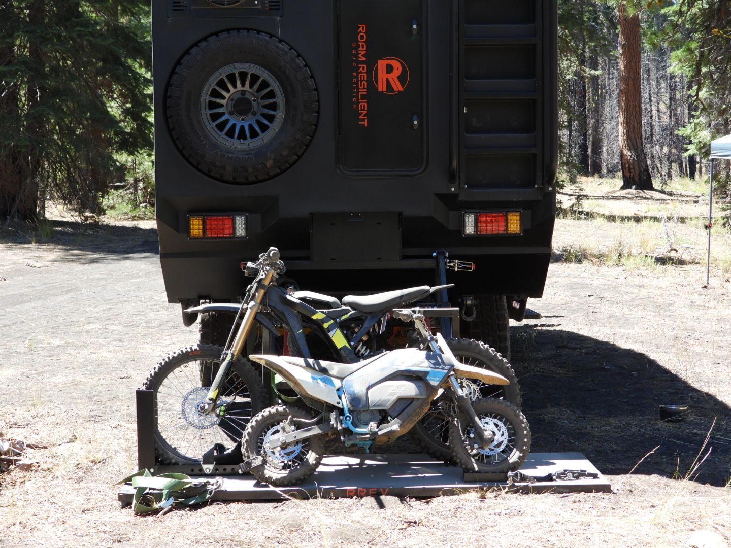 Two electric dirt bikes strapped to the rear carrier below the spare tire