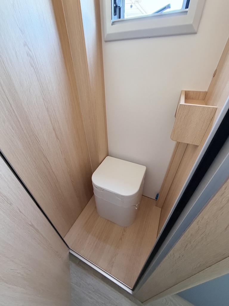 Portable toilet tucked into an oak-lined bathroom corner below a small opening window