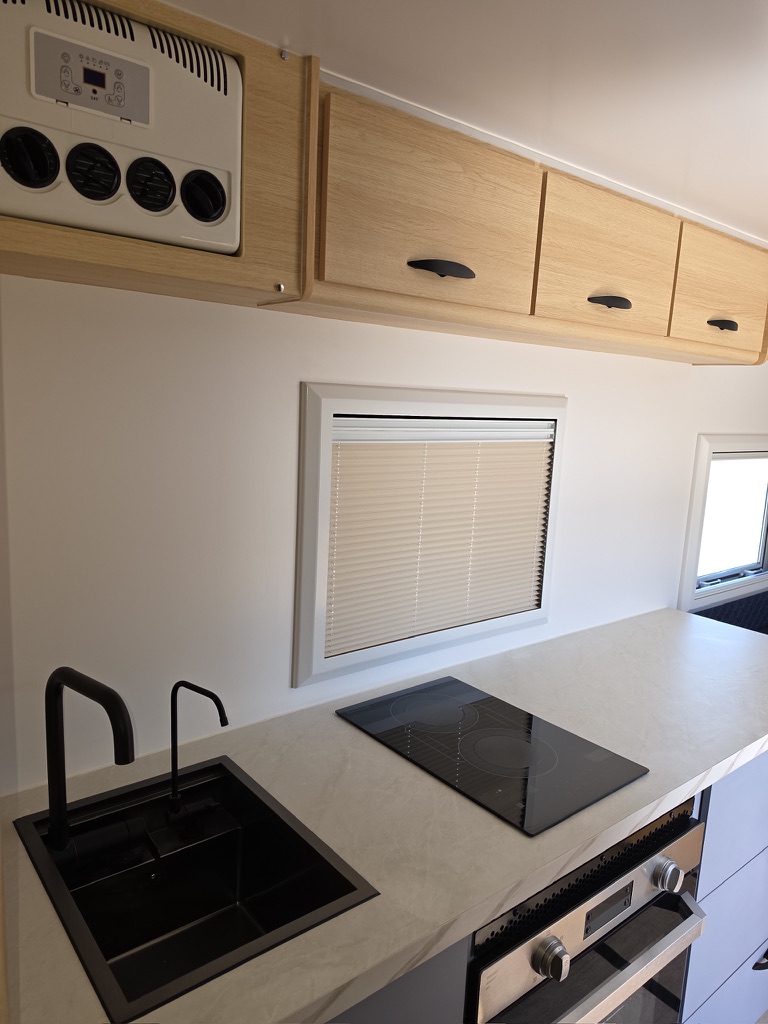 Black sink and induction hob beneath oak overhead lockers, with the air conditioner head and window blinds