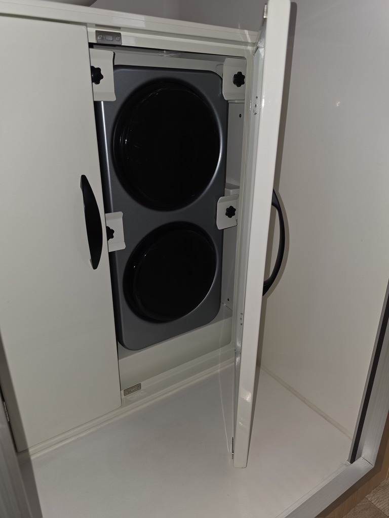 Two-burner glass cooktop stowed upright in a galley cabinet with quick-release retaining knobs